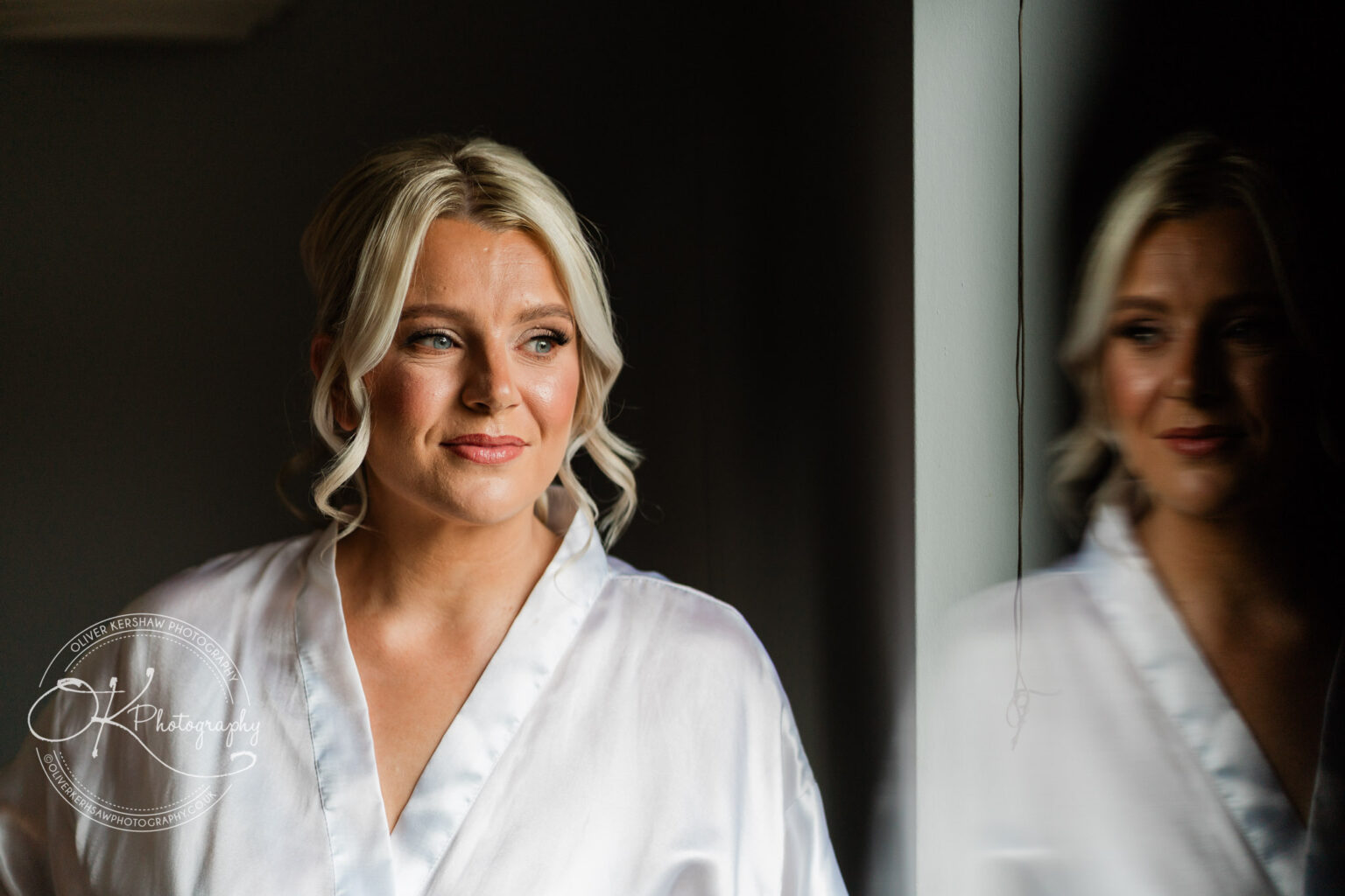 Woman in a white robe smiling softly, with her reflection visible in a dark mirror.