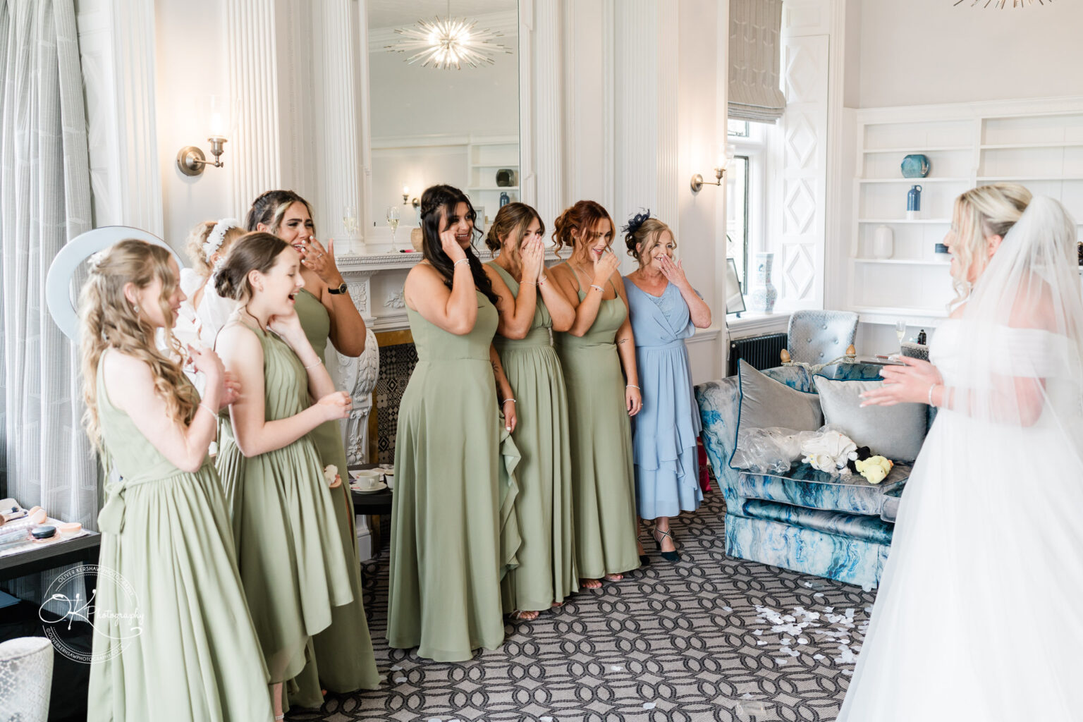 A group of bridesmaids in olive green dresses, with expressions of surprise and joy, gather around a bride in a white dress and veil. The scene takes place in a stylish room with elegant decor, including a blue sofa and a decorative fireplace.