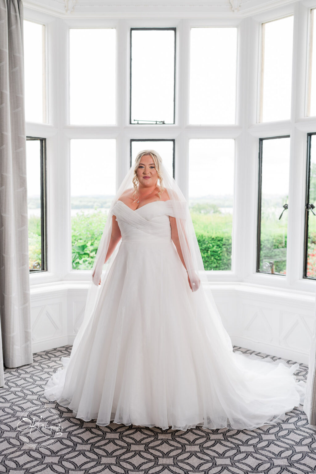 A bride in a white wedding gown with an off-the-shoulder neckline stands gracefully in a bright room, surrounded by large windows. She has long, wavy hair and wears a sheer veil. The lush green landscape is visible through the windows, adding a serene backdrop to the scene.