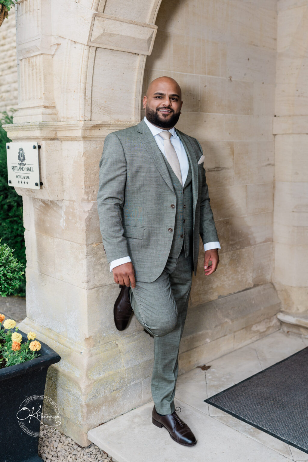 A man in a light grey suit with a waistcoat and a white shirt leans against a stone wall, smiling. He has a bald head and a neatly trimmed beard. In the background, there's a sign for Rutland Hall Hotel & Spa and a flower pot with marigolds.