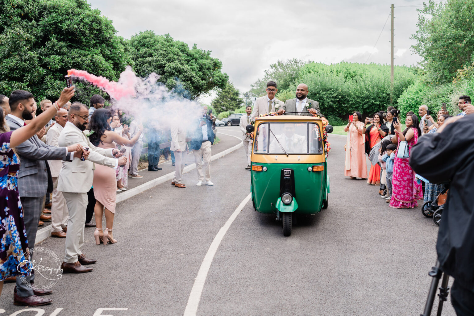 A vibrant outdoor scene featuring two men in suits riding in a decorated green and yellow auto rickshaw. Surrounding them are celebration attendees holding sparklers and taking photos, with trees and a cloudy sky in the background.