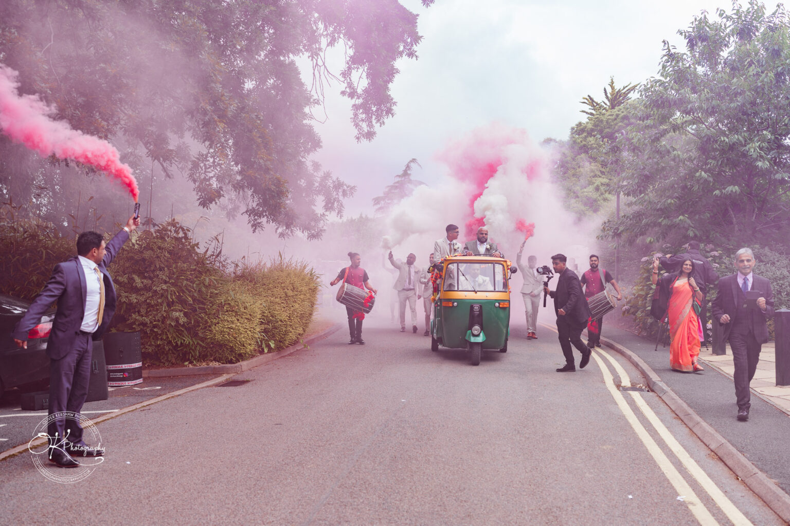 A vibrant scene of a wedding procession featuring a decorated auto rickshaw surrounded by jubilant guests. Smoke flares release pink smoke in the air, while individuals in suits and traditional attire celebrate, some playing drums and others capturing the moment on camera. Lush greenery borders the roadside.