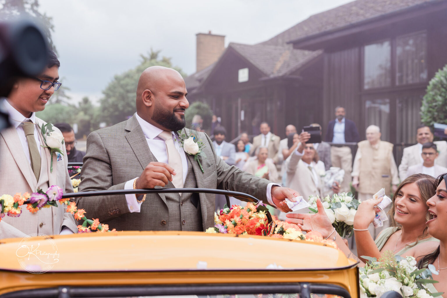 A groom in a light grey suit hands money to a guest while sitting in a yellow decorative car surrounded by wedding guests, who are smiling and holding flowers.