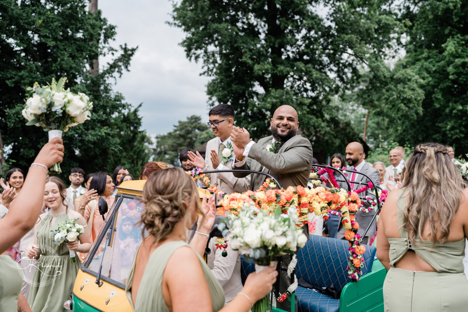 A joyful scene with a smiling man in a grey suit riding in a decorated auto rickshaw, surrounded by guests celebrating, holding flowers, and clapping. Lush green trees are in the background, under a slightly cloudy sky.