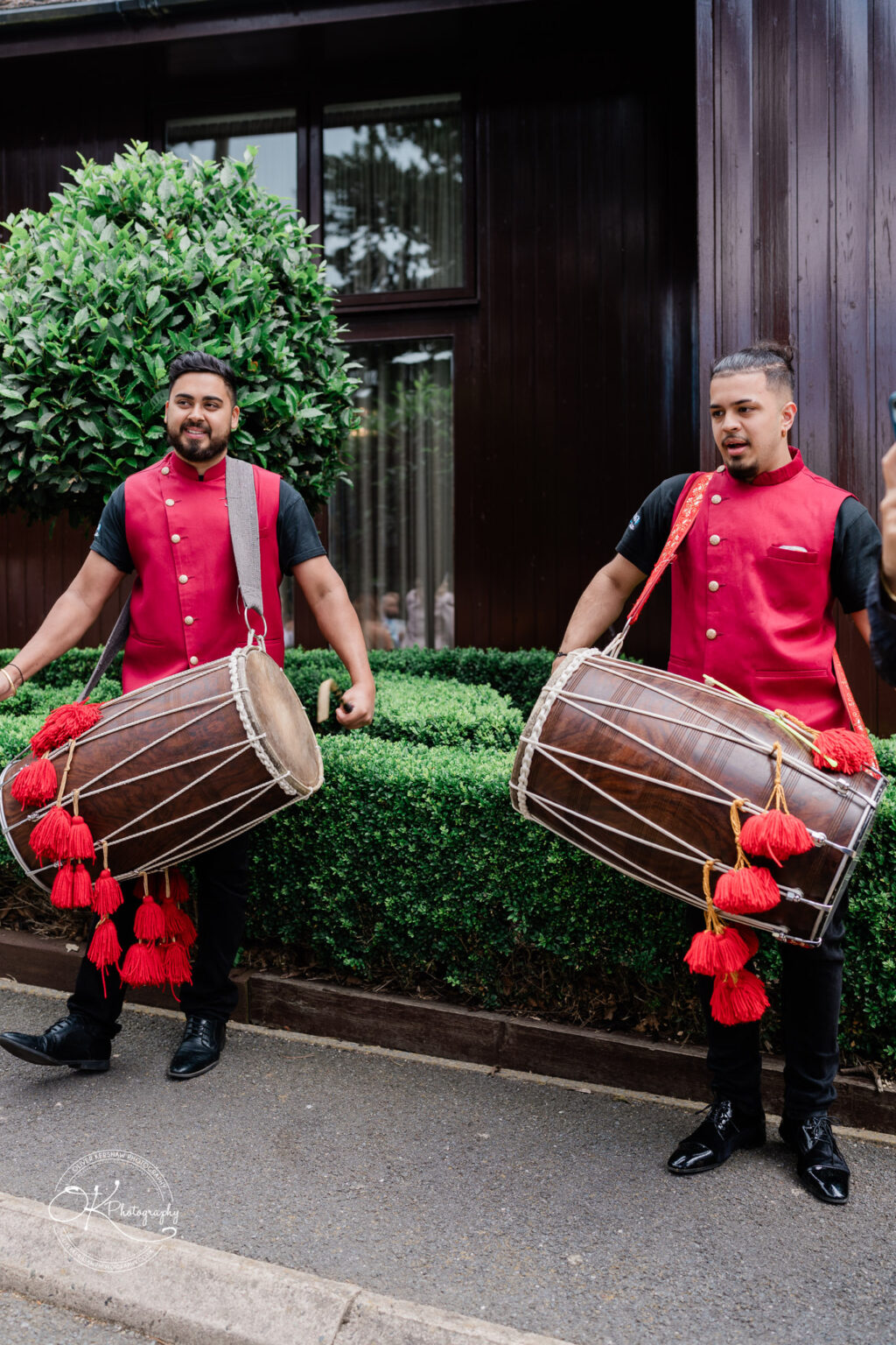 Two men in traditional red and black outfits are playing large drums decorated with red tassels. They are standing outdoors near a neatly trimmed hedge and a dark wooden building.