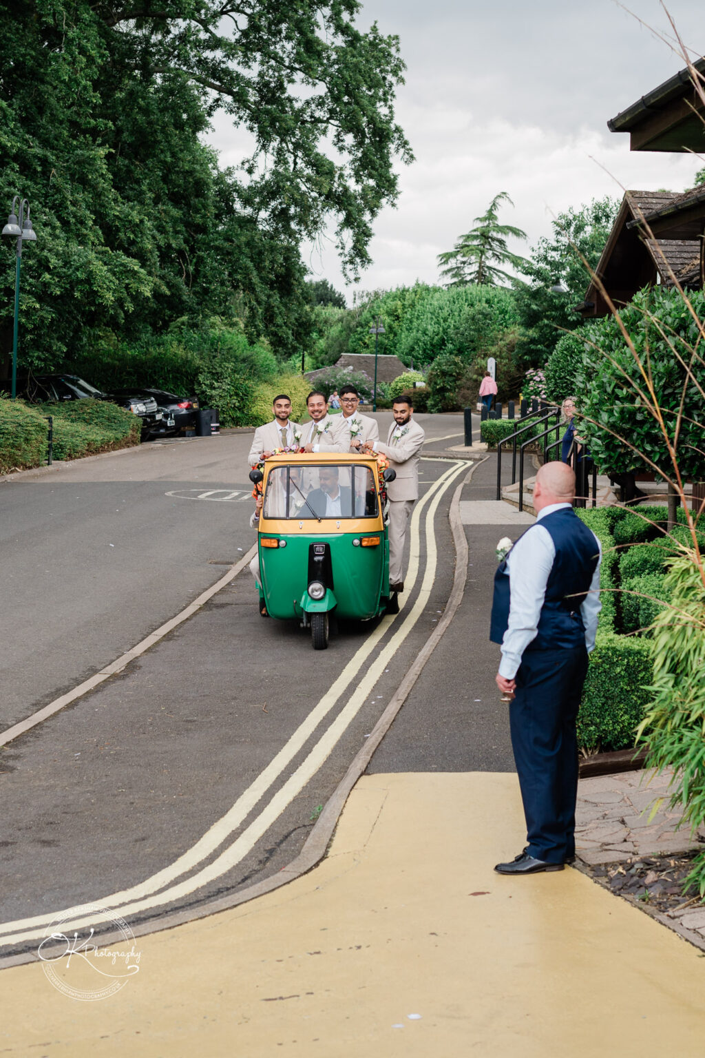 A group of four men in light-coloured suits and one man in a dark suit are interacting by a vibrant green and yellow auto rickshaw on a scenic path with trees and greenery in the background.
