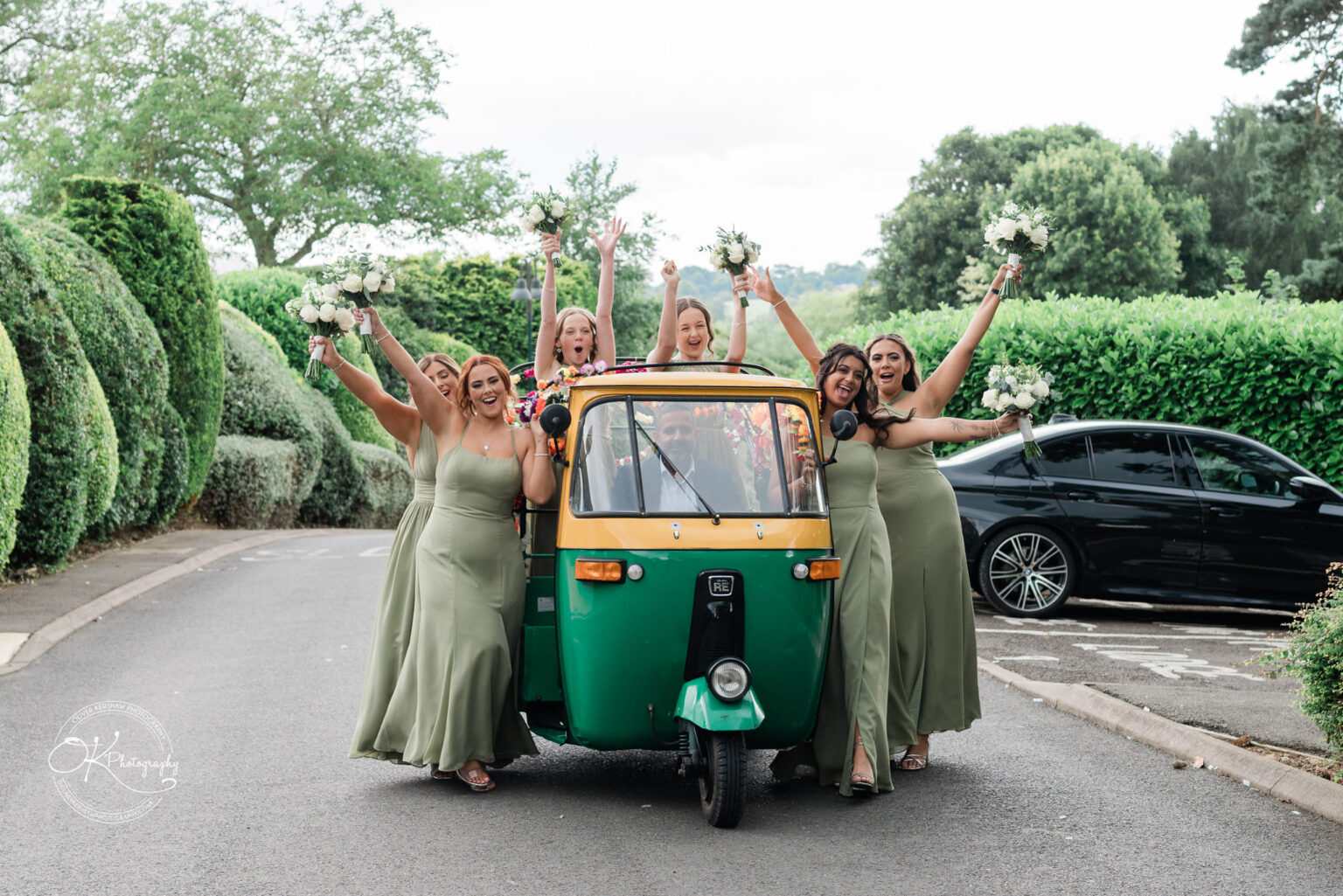 A group of six women in matching green dresses celebrate around a colourful tuk-tuk, holding floral bouquets and posing joyfully. In the driver's seat, a man smiles as they all cheer, set against a backdrop of neatly trimmed hedges and a black car parked nearby.