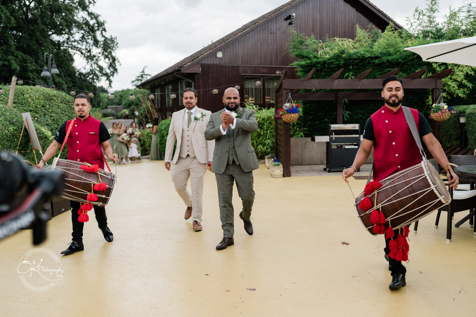 A groom in a light-coloured suit walks joyfully down a pathway, flanked by two drummers in red and black attire playing traditional drums. In the background, a wedding party in green dresses is visible, with a rustic venue and greenery surrounding the scene.