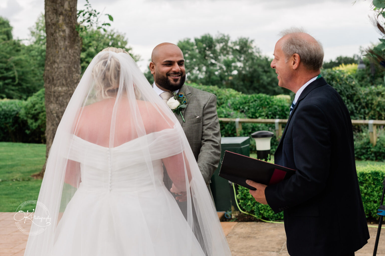 A bride in a white off-the-shoulder wedding gown and veil stands holding hands with a groom in a grey suit, smiling at an officiant who is holding a book. The setting is an outdoor garden with greenery in the background.