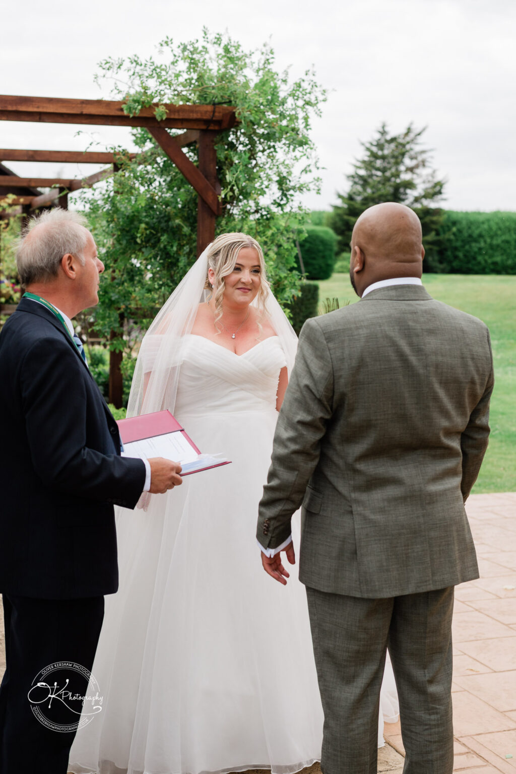 A bride in a white gown and veil stands smiling in front of a groom in a grey suit. They are joined by a man officiating the wedding, holding a binder. The backdrop features greenery and a cloudy sky.