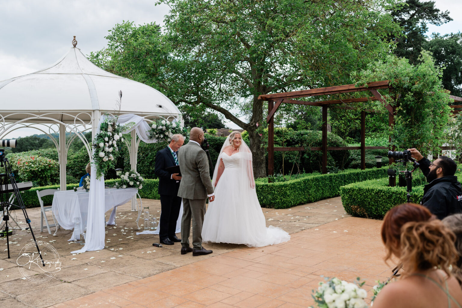 A bride in a white gown with a veil stands next to a groom during a wedding ceremony under a decorated gazebo. A person in formal attire officiates, while a photographer captures the moment. The setting features greenery and floral decorations.