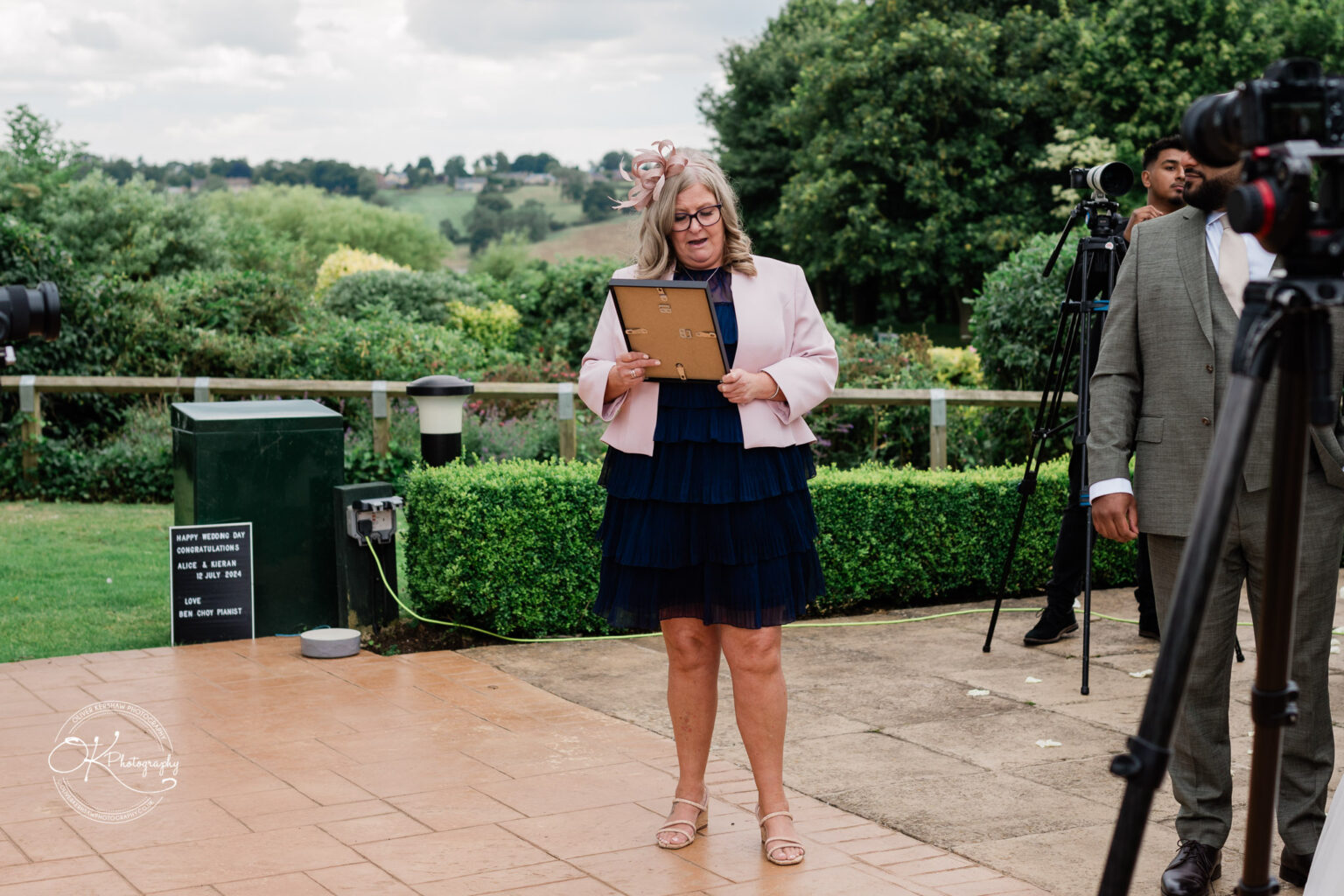 A woman with long hair, wearing glasses and a pink jacket, is reading from a clipboard at an outdoor wedding ceremony. There are photographers and guests in the background, and a sign nearby congratulates the couple.