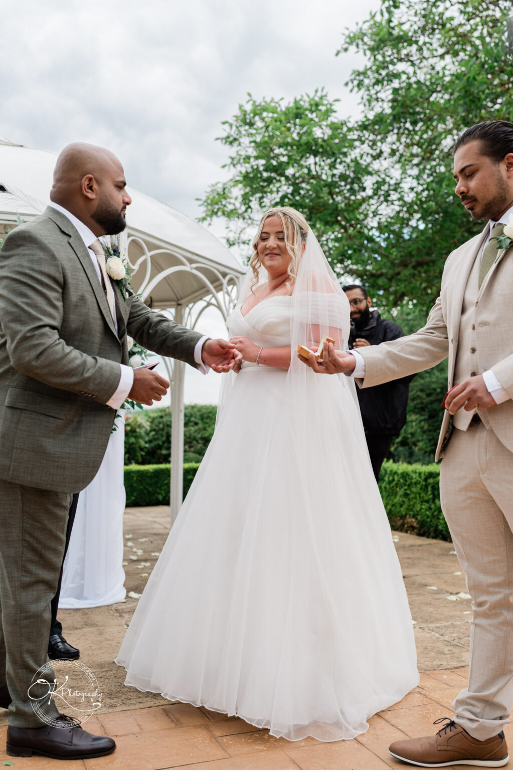 A bride in a white wedding dress and veil smiles as she holds hands with a groom in a grey suit during a wedding ceremony, with a man in a beige suit presenting a ring. The setting features greenery and a gazebo.