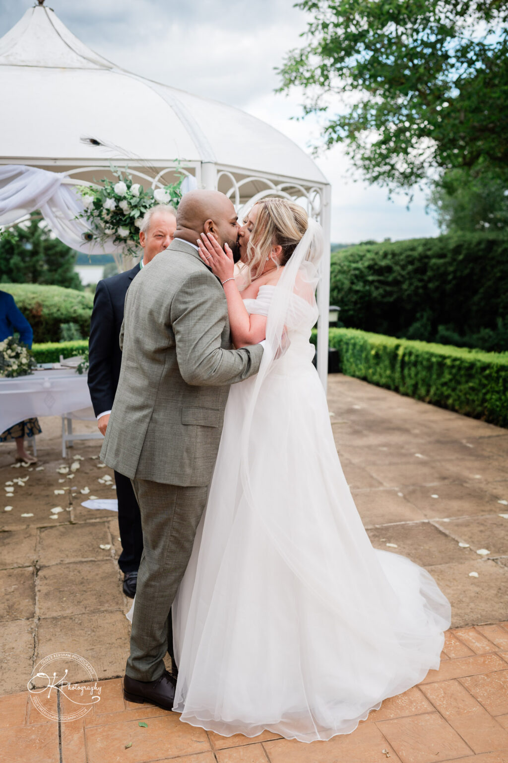 A couple in wedding attire is sharing a kiss under a gazebo, with a floral arrangement in the background. The bride is wearing a white strapless dress and veil, while the groom is in a grey suit. An officiant is visible in the background. Rose petals are scattered on the ground.