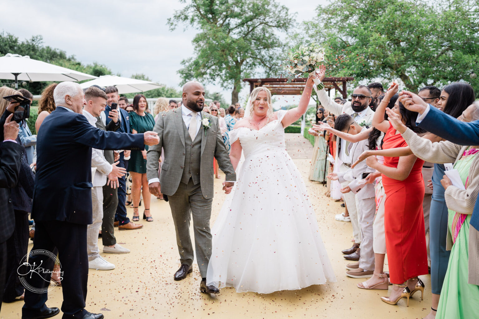 A bride and groom walk hand in hand down an aisle, smiling brightly as guests throw confetti. The bride wears a white off-the-shoulder gown and holds a bouquet, while the groom is dressed in a grey suit. Guests are dressed in various attire, with some clapping and others taking photos in the background.
