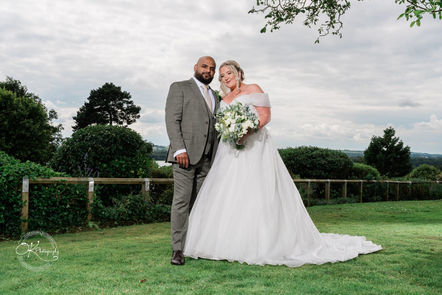 A bride wearing a white off-shoulder wedding dress stands beside a groom in a grey suit on a grassy area, with greenery and a cloudy sky in the background. The bride is holding a bouquet of white flowers.