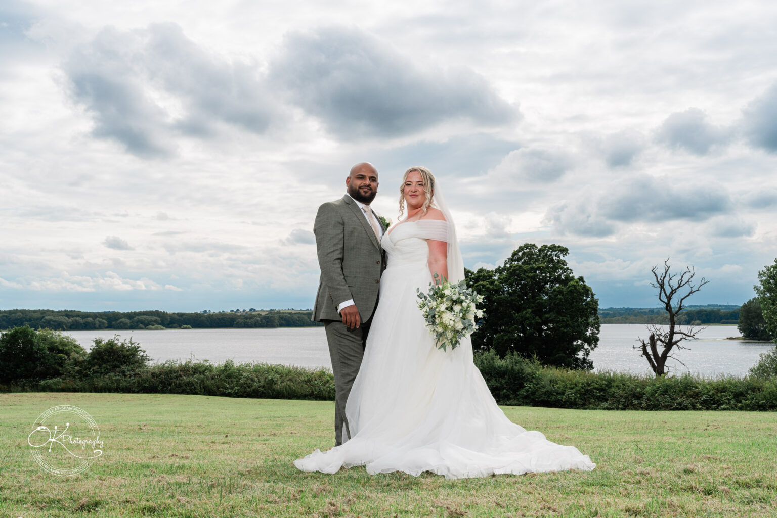 A couple stands together on a grassy hill with a scenic lake in the background. The man is dressed in a grey suit, while the woman wears a white wedding gown and holds a bouquet of flowers. The sky is partially cloudy.