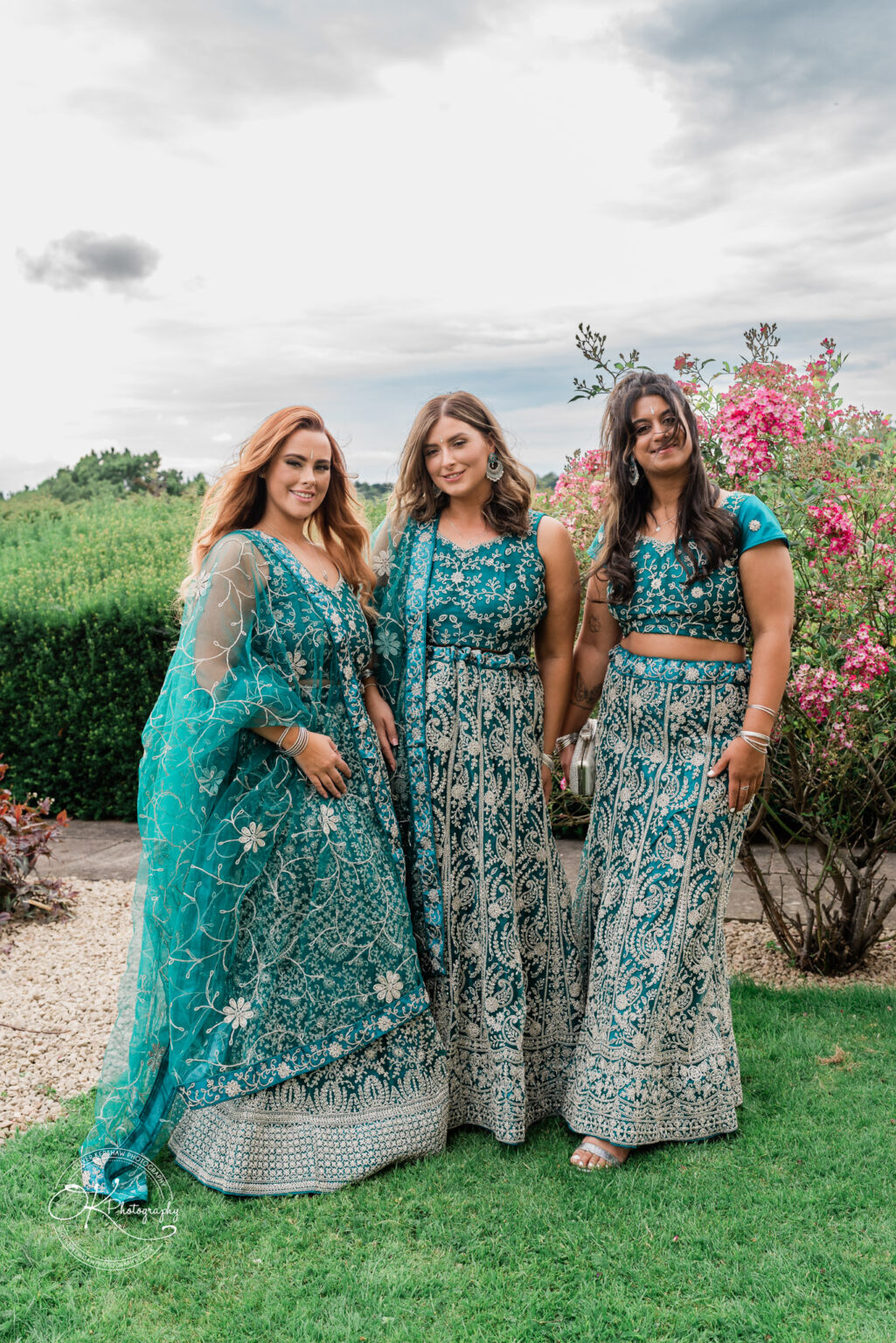 Three women stand together in a garden, dressed in traditional teal-coloured outfits with intricate silver embroidery. They are smiling, with floral decorations and greenery in the background.