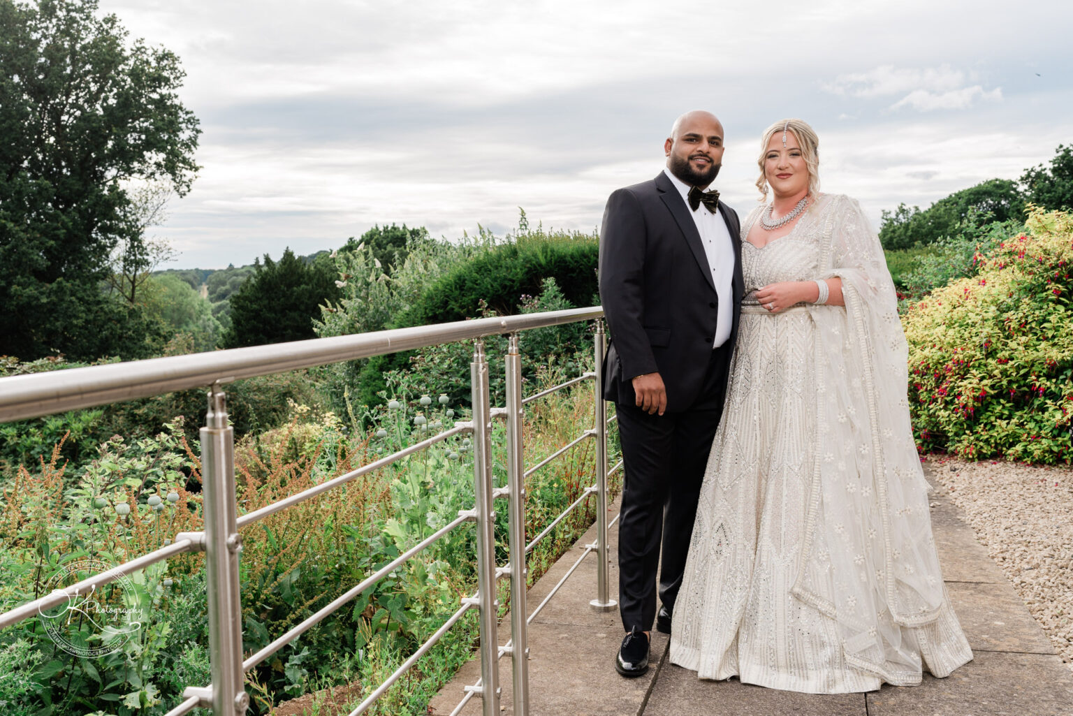A couple stands together on a balcony, dressed elegantly for a formal event. The man wears a black tuxedo with a bow tie, while the woman is in a beautifully detailed light-coloured gown with intricate embellishments. They are surrounded by lush greenery and flowers, with a cloudy sky in the background.