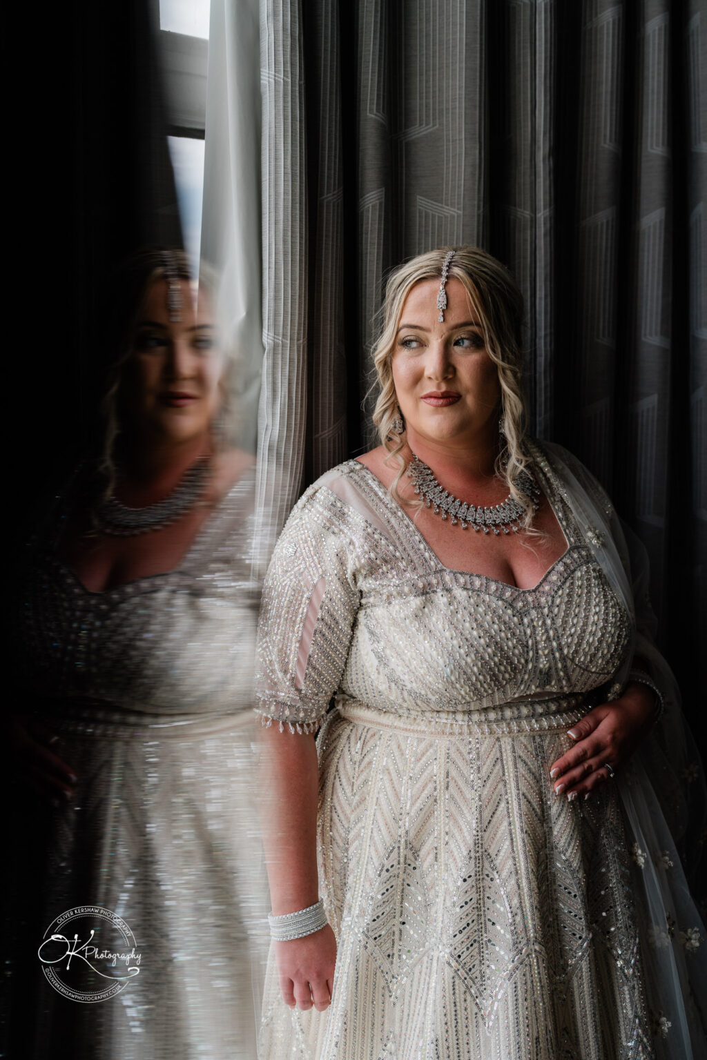 A woman in an elegant, intricately beaded dress stands by a window, looking thoughtfully to the side. Her hair is styled in loose curls, and she wears a decorative headpiece and layered necklace. A soft reflection of her image is visible on the window.