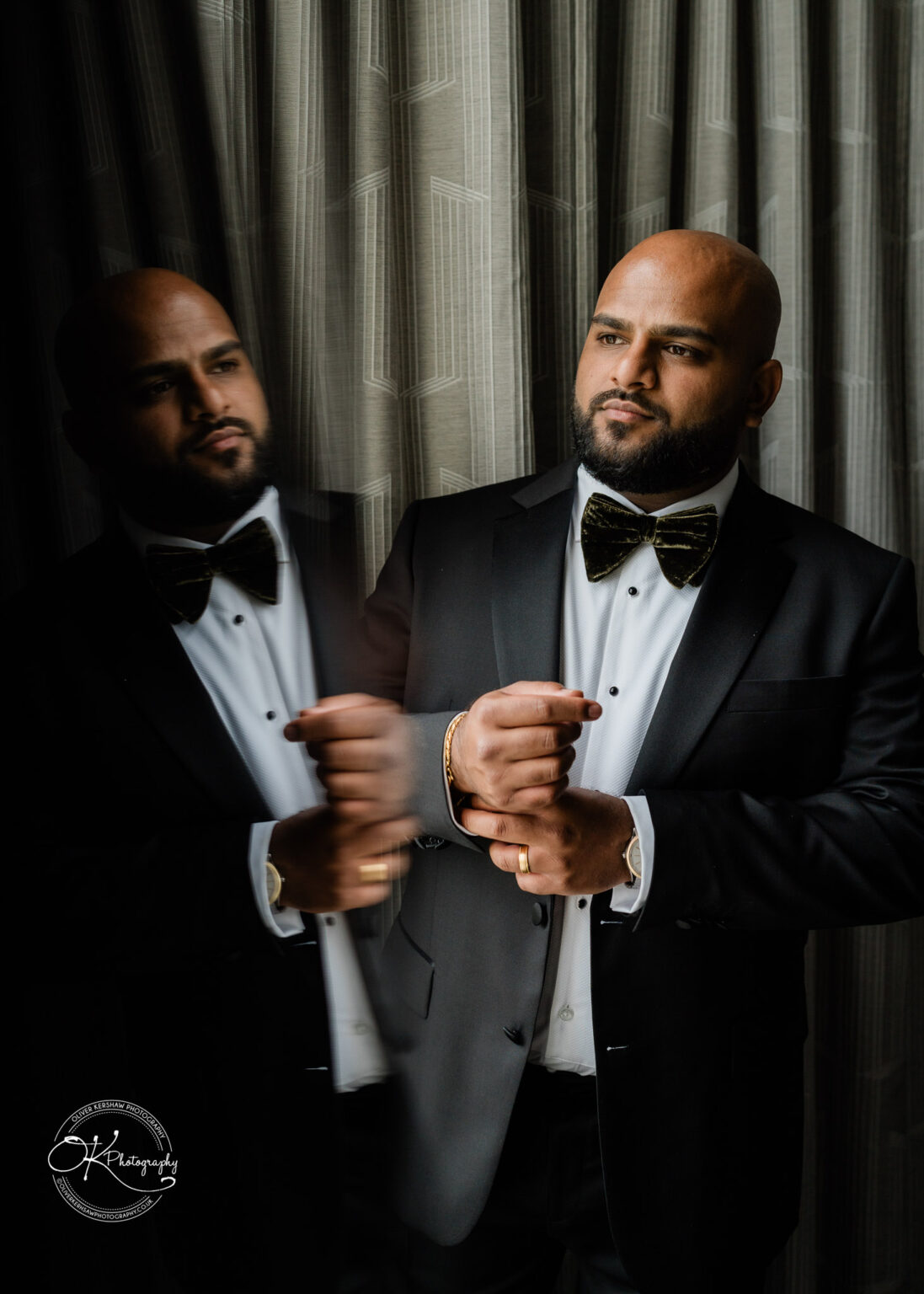 A well-dressed man in a tuxedo and bow tie adjusts his sleeve cuffs in front of a curtain, with a reflection visible beside him. He has a bald head and a beard, exuding a confident demeanor.