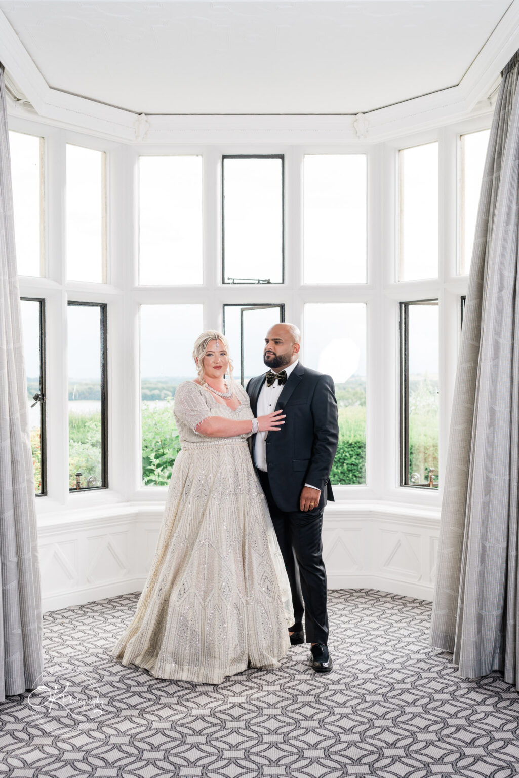 A couple stands together in a bright room with large windows, surrounded by green scenery. The woman is wearing a sparkling, elaborately beaded gown, while the man is dressed in a classic black tuxedo with a bow tie.