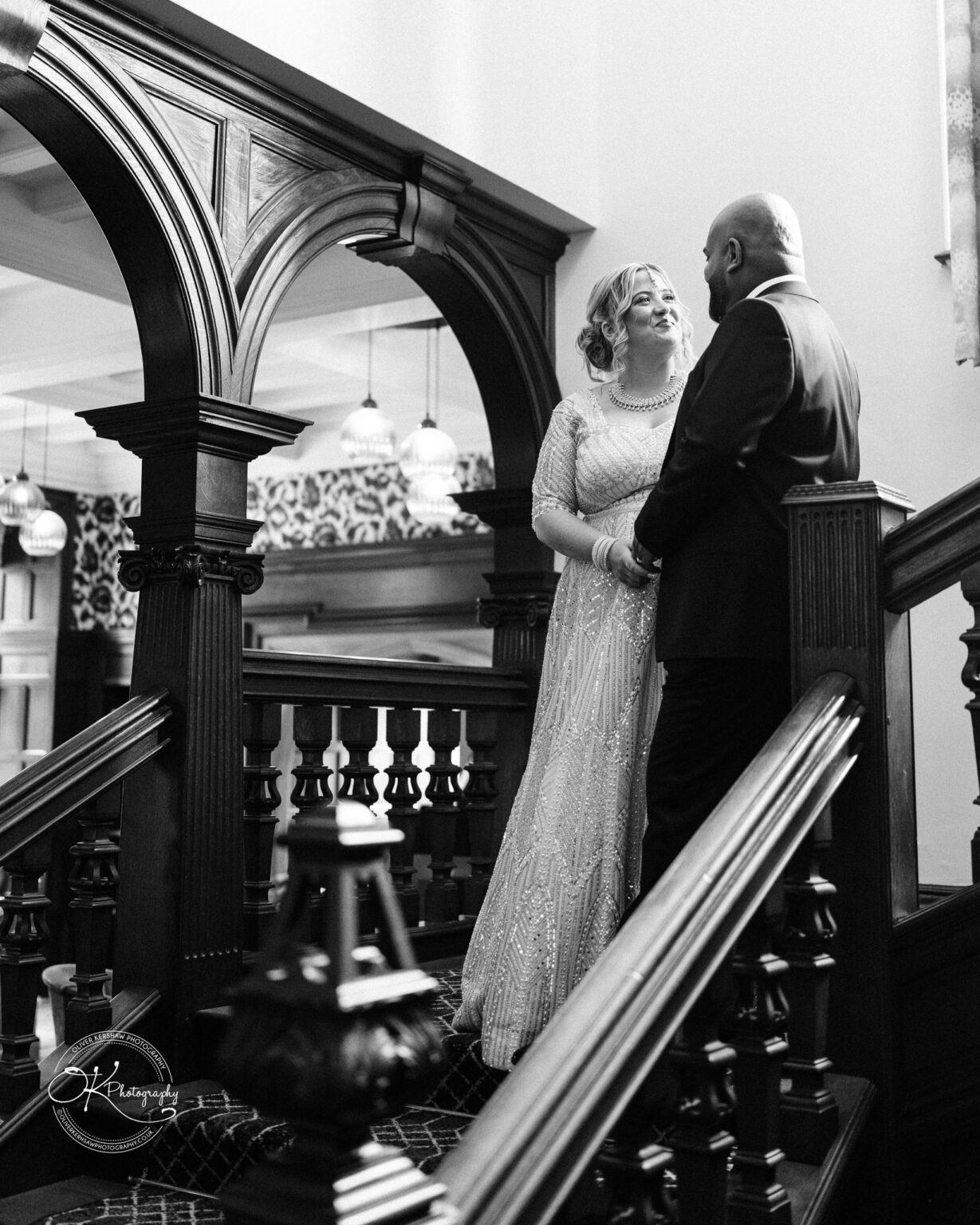 A couple stands on a staircase, gazing at each other lovingly. The woman wears a sparkling, intricate gown, and the man is dressed in a suit. The setting features elegant wooden architecture and soft lighting.