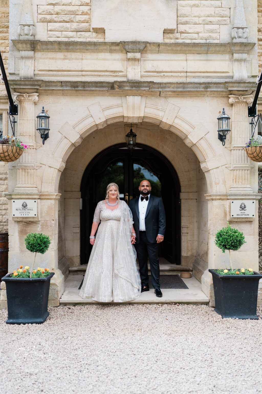 A couple stands hand in hand in front of an elegant stone building entrance, the woman wearing a beautifully beaded light grey gown and the man in a classic black tuxedo. Flower baskets adorn the entrance, and there are two potted plants on either side of them.