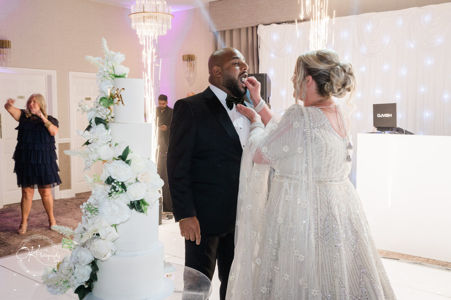 A bride in a sparkling white dress playfully feeds a piece of cake to a groom in a black tuxedo, with a large decorated wedding cake nearby. In the background, an excited guest is clapping, and there are sparklers providing a festive atmosphere.