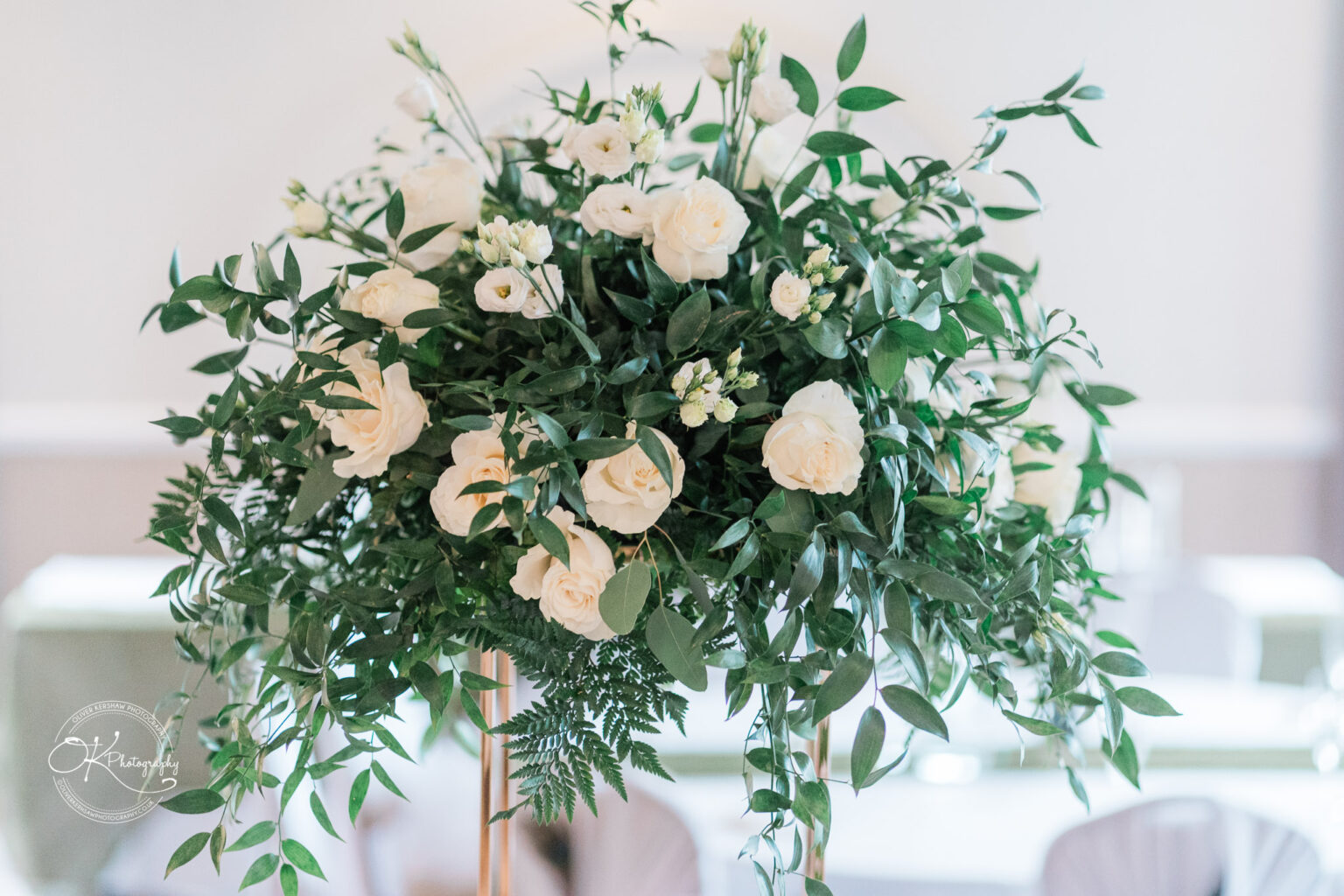 A lush floral arrangement featuring white roses and various greenery, atop a tall stand, with a softly blurred background.