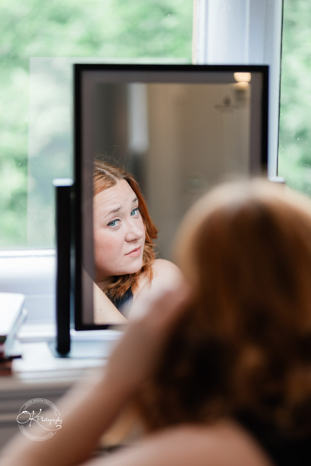 A woman with auburn hair and blue eyes is sitting in front of a mirror, looking at her reflection with a thoughtful expression. The background features greenery visible through a window.