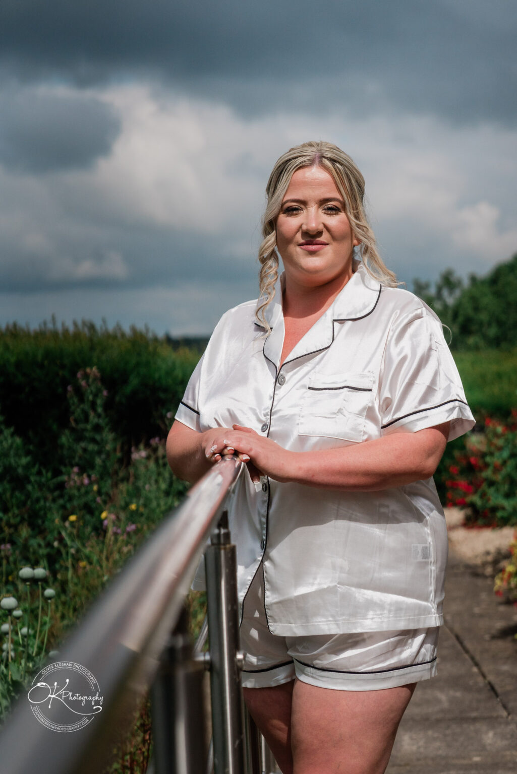 A woman in silk pyjamas stands by a rail in a garden, with green bushes and flowers in the background. The sky is partly cloudy.