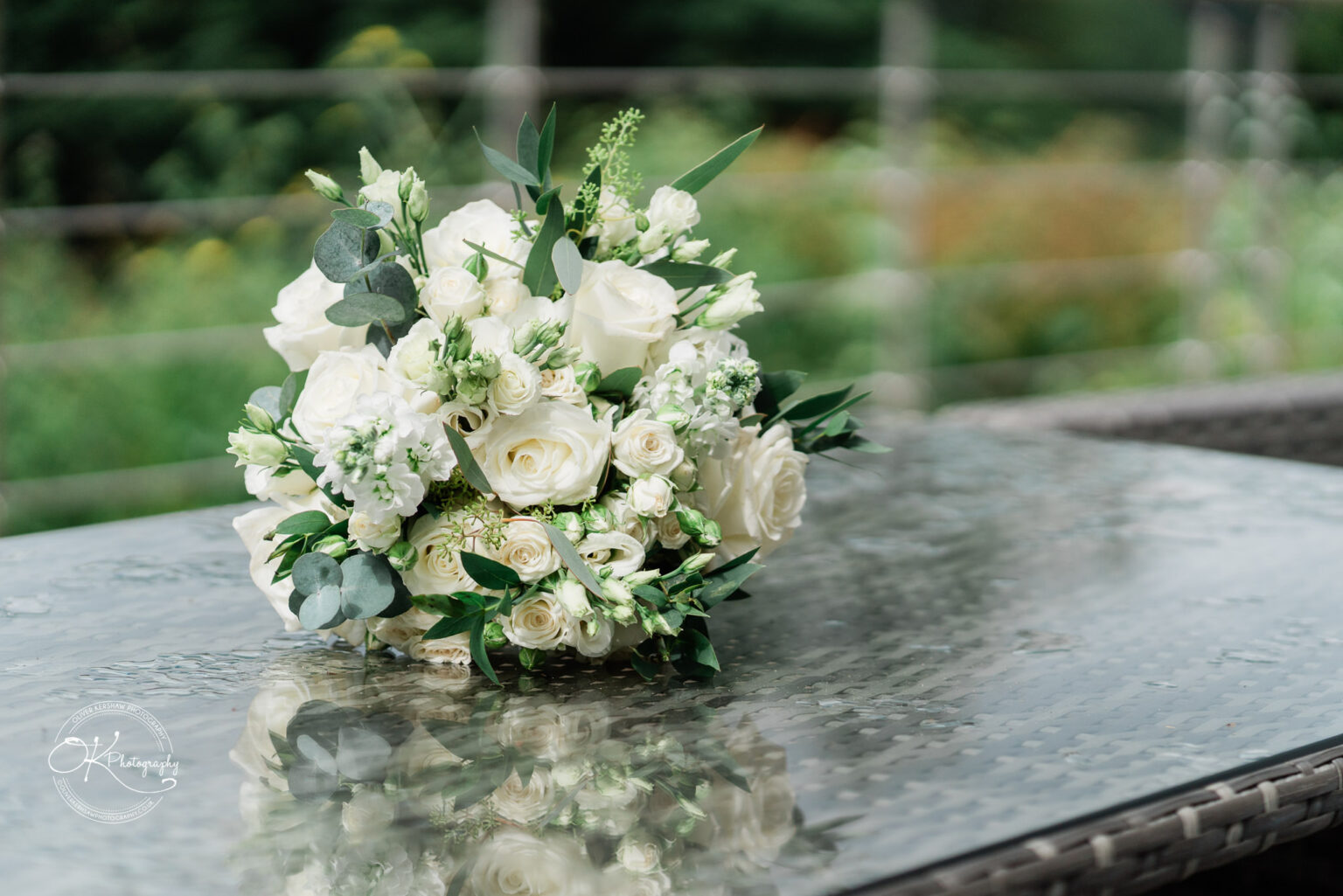 A beautifully arranged bouquet of white roses and greenery placed on a glass table, with a blurred natural background.