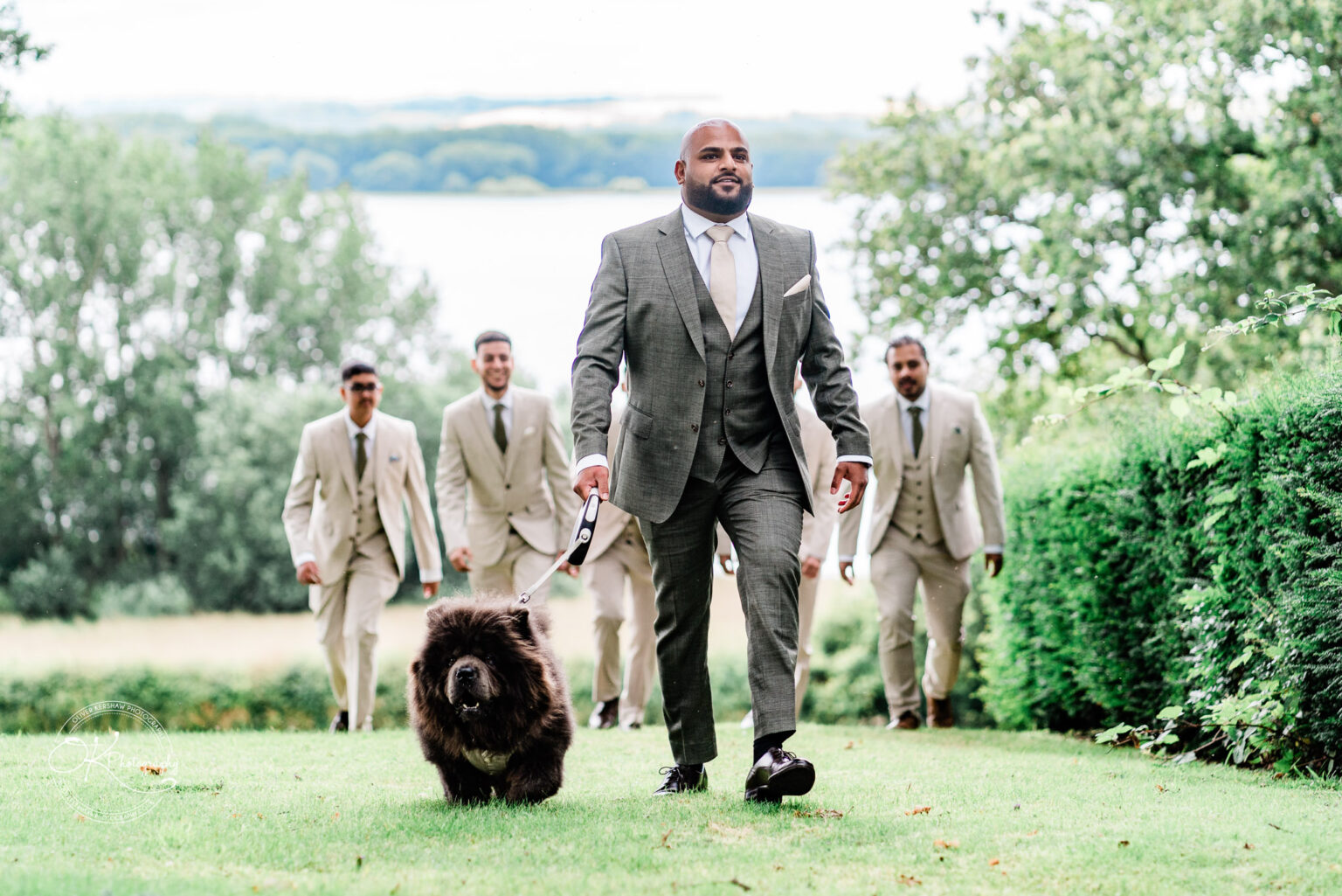 A man in a grey suit walks confidently on grass, holding a leash attached to a fluffy brown dog, while several men in beige suits follow behind them, with trees and a lake visible in the background.