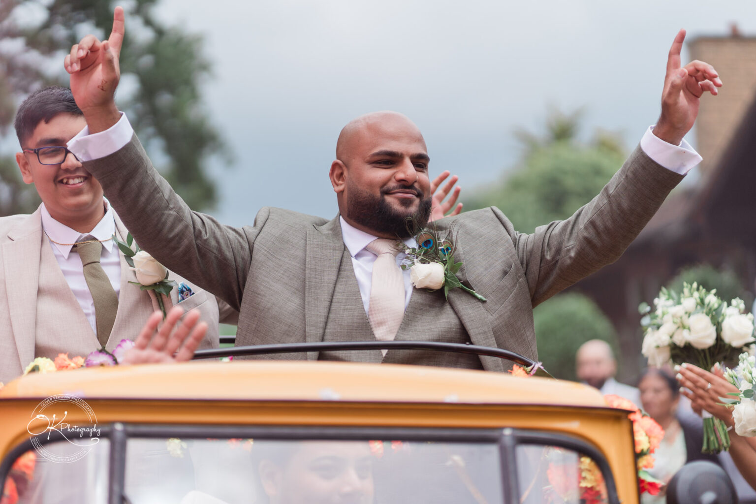 A joyful man in a grey suit, smiling and raising his arms in celebration, rides in a yellow decorated vehicle with flowers, while another man in a light suit stands beside him, both surrounded by cheering guests.