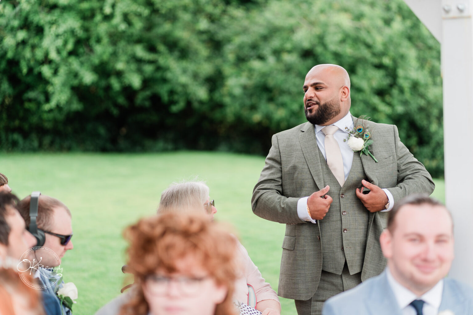 A well-dressed man in a grey suit with a light-coloured tie is animatedly speaking at a wedding ceremony, while seated guests listen attentively. Some guests have headphones on, and greenery can be seen in the background.
