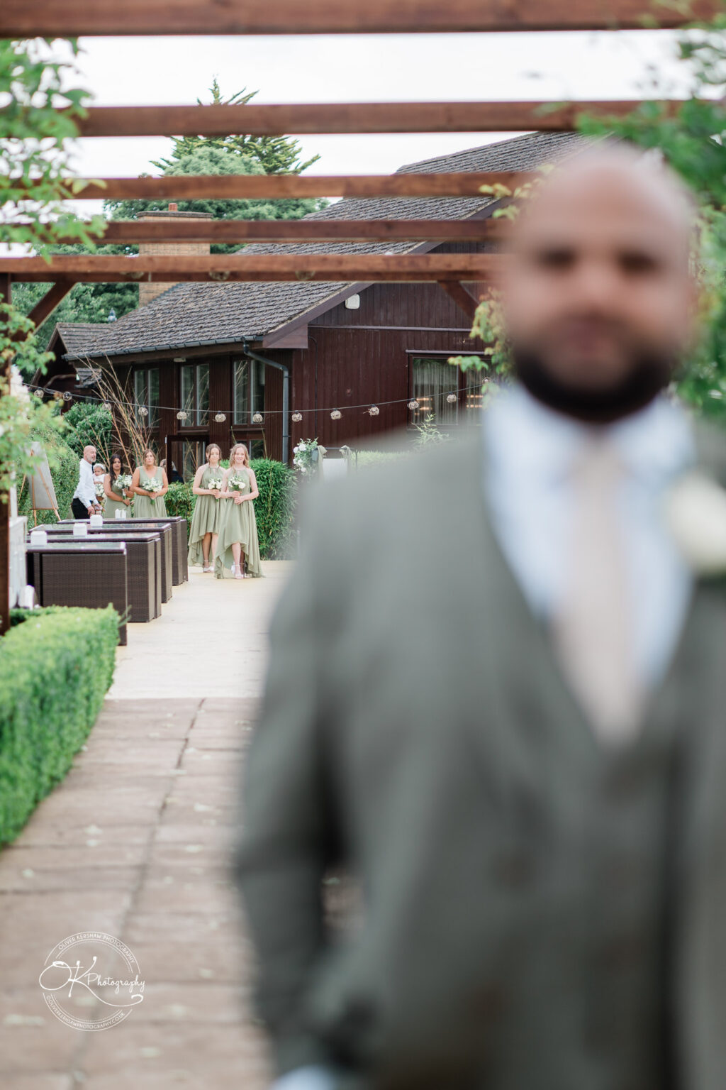 A blurred foreground figure in formal attire stands in focus, while in the background, a group of women in green dresses walks along a path towards a wooden building, surrounded by greenery.
