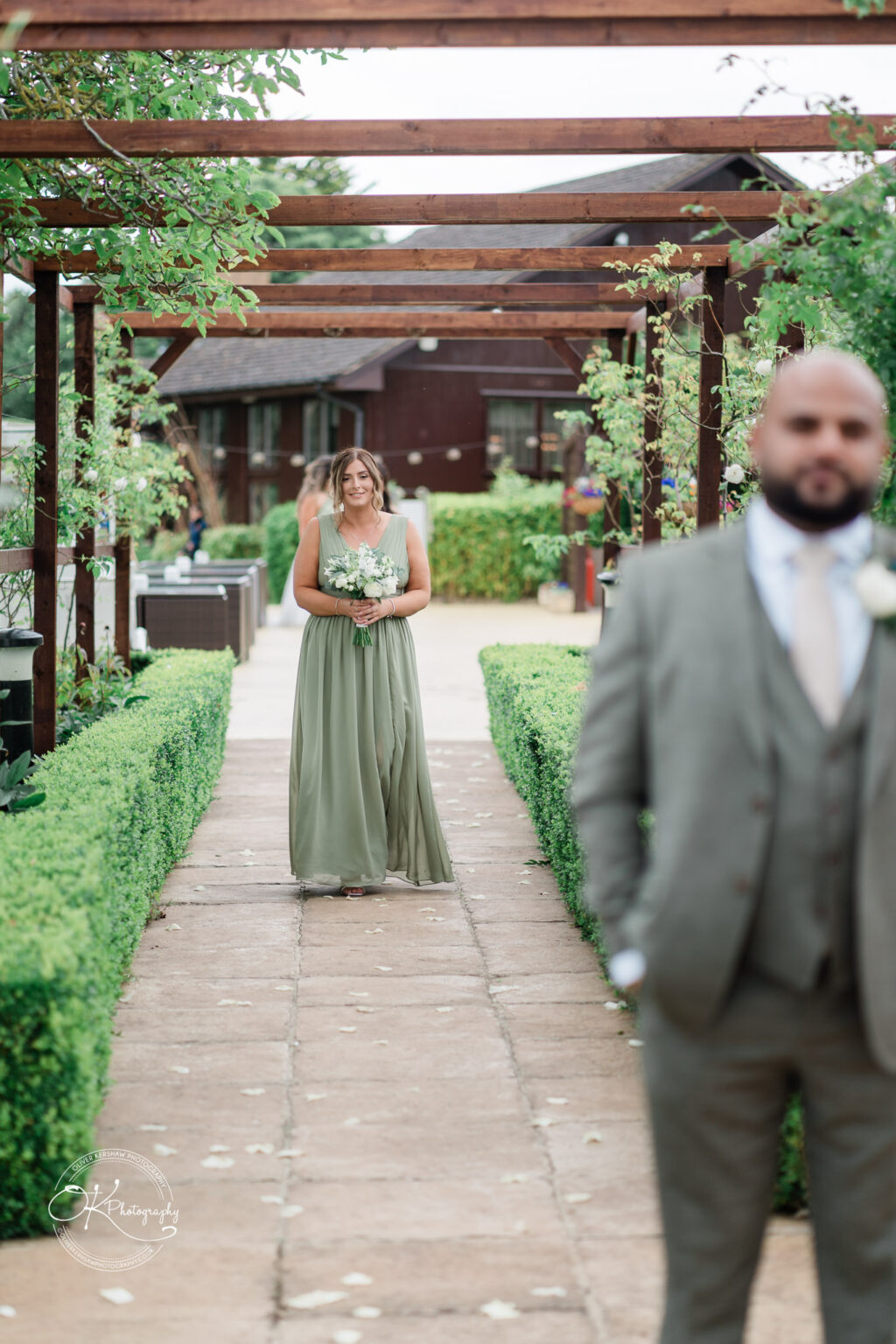 A woman in a flowing green dress holding a bouquet walks down a garden path lined with hedges, while a man in a grey suit stands slightly blurred in the foreground.