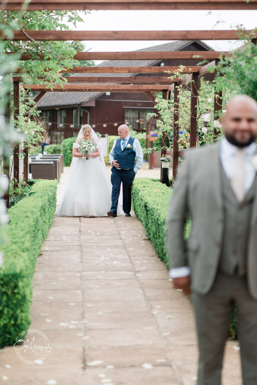 A bride in a white gown and veil walks down a garden path with her father, who is dressed in a blue waistcoat. They are surrounded by neatly trimmed hedges and wooden pergolas adorned with greenery. A blurred figure of a man in a grey suit stands in the foreground.