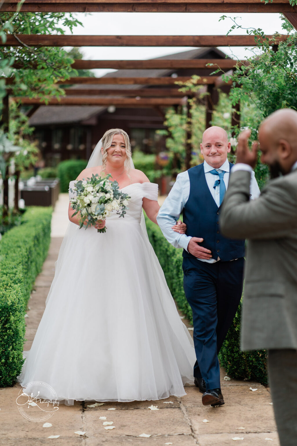 A bride in a white off-the-shoulder wedding gown walks down an outdoor path lined with greenery, holding a bouquet of flowers, accompanied by a man in a blue vest and trousers, while another man in a suit waves from the side.