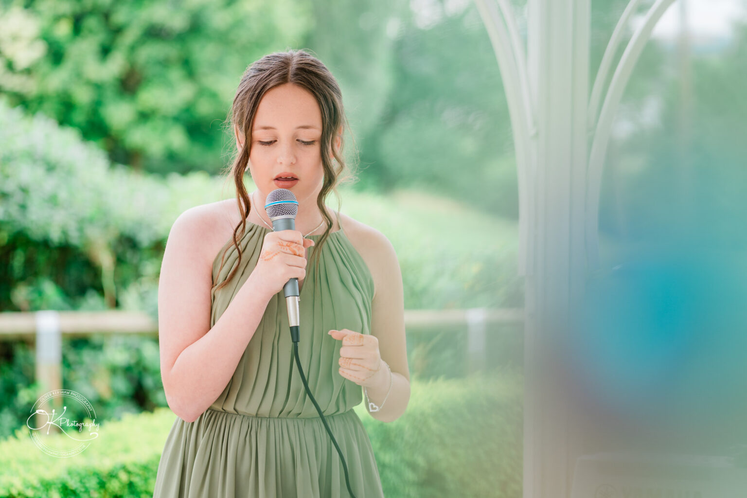 A young woman in a green dress sings into a microphone, with a soft-focused background of greenery.