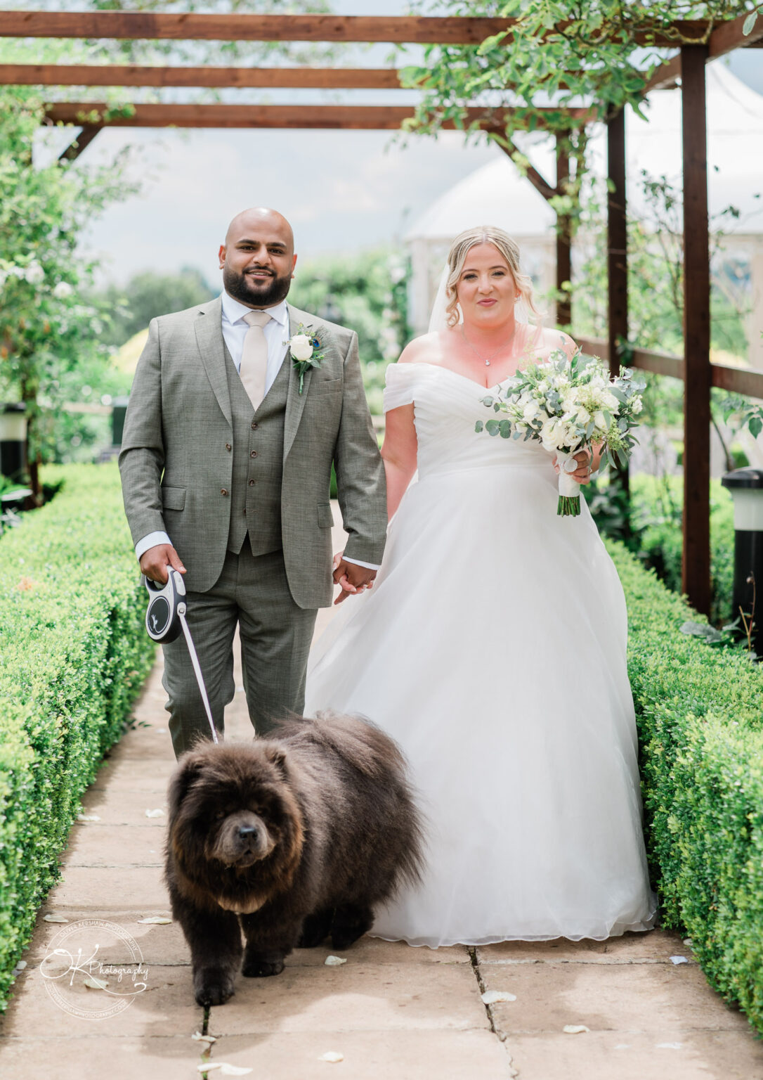 A bride in a white dress and a groom in a grey suit hold hands while walking down a pathway lined with greenery, accompanied by a large, fluffy dog on a leash.