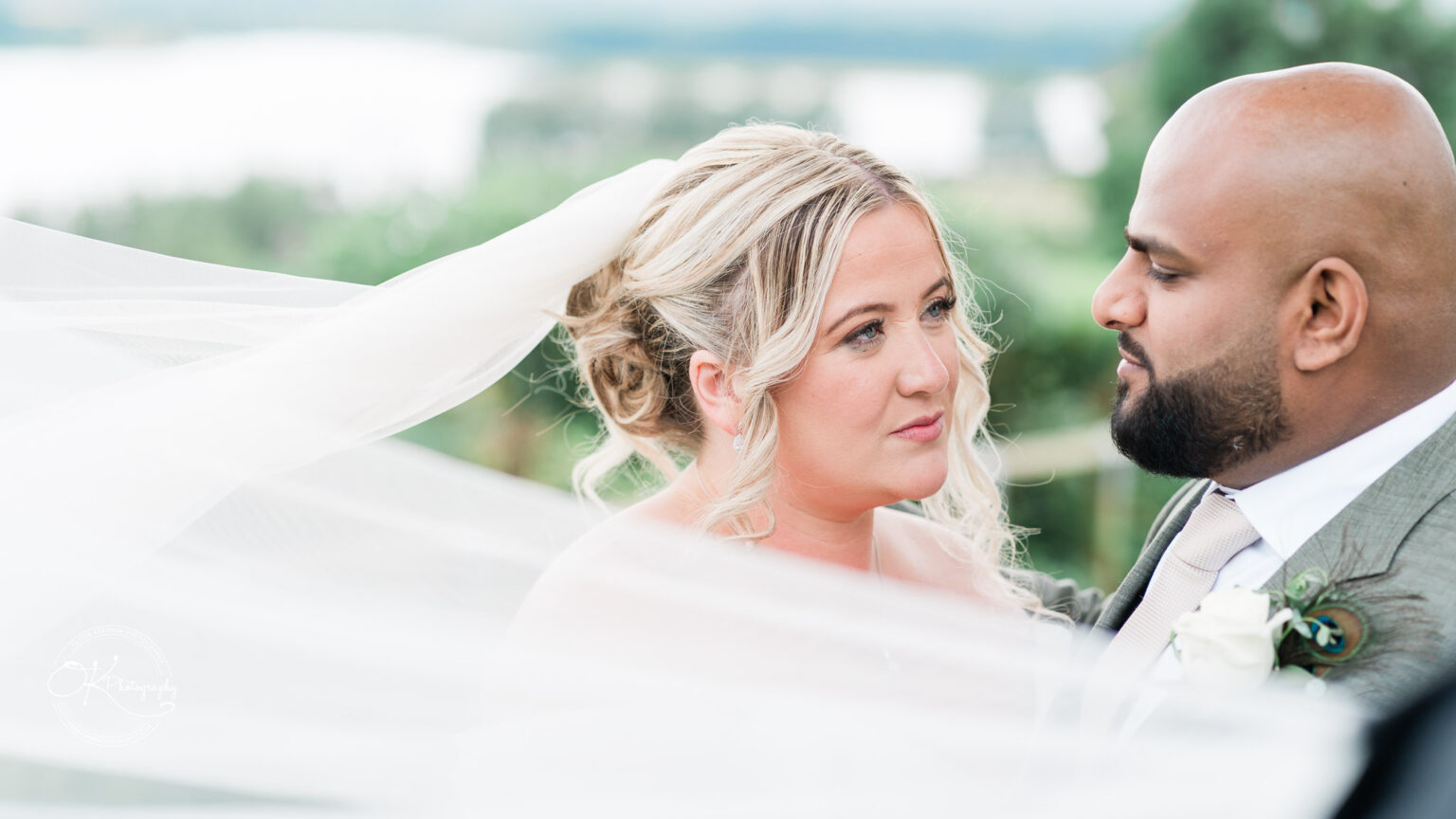 A bride with wavy blonde hair and a veil gazes at her partner, who has a shaved head and beard, as they share an intimate moment outdoors with blurred greenery and a body of water in the background.