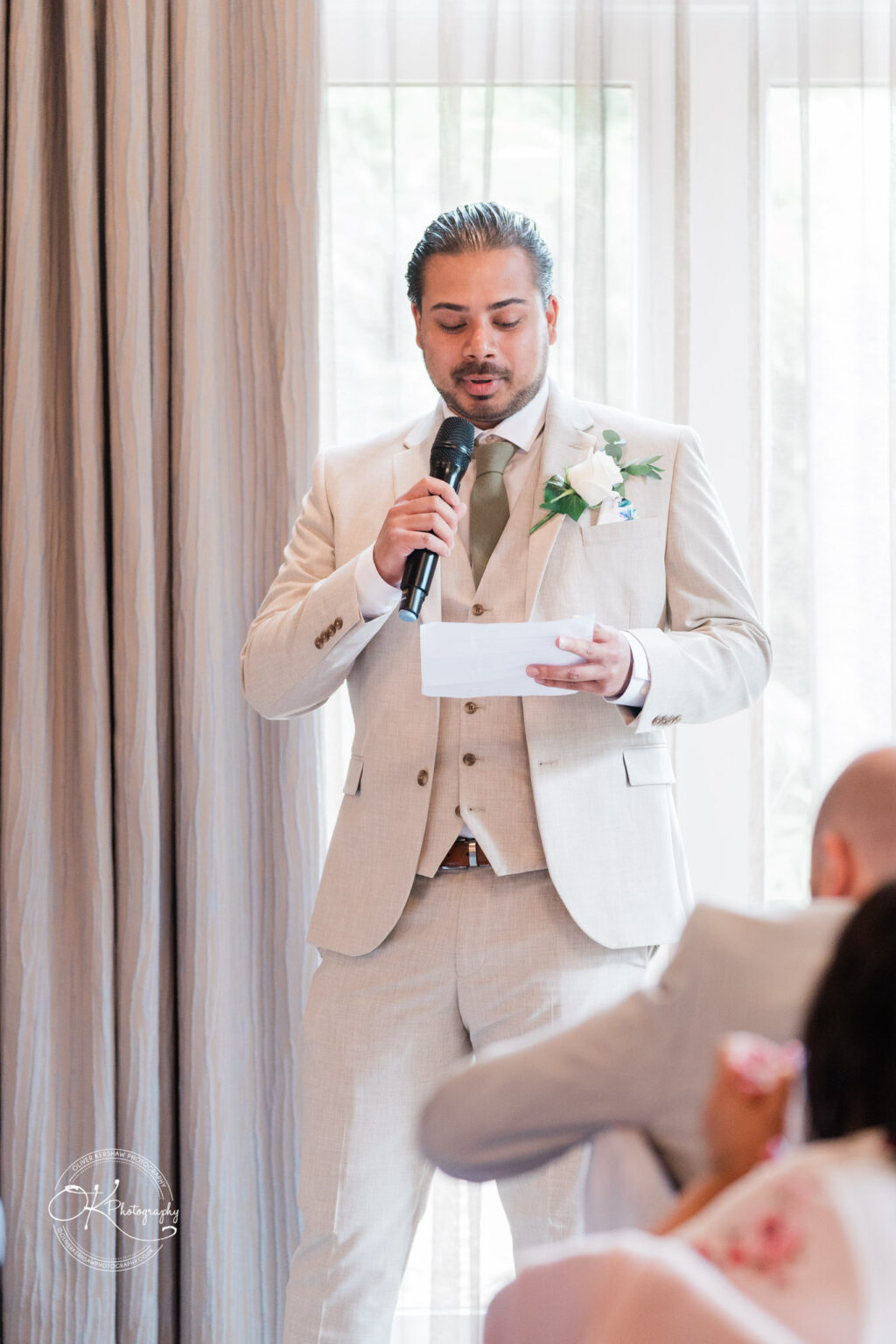 A man in a light beige suit is speaking into a microphone while holding a piece of paper, standing in front of large curtains. He has a white flower pinned to his lapel and appears to be addressing an audience seated in front of him.