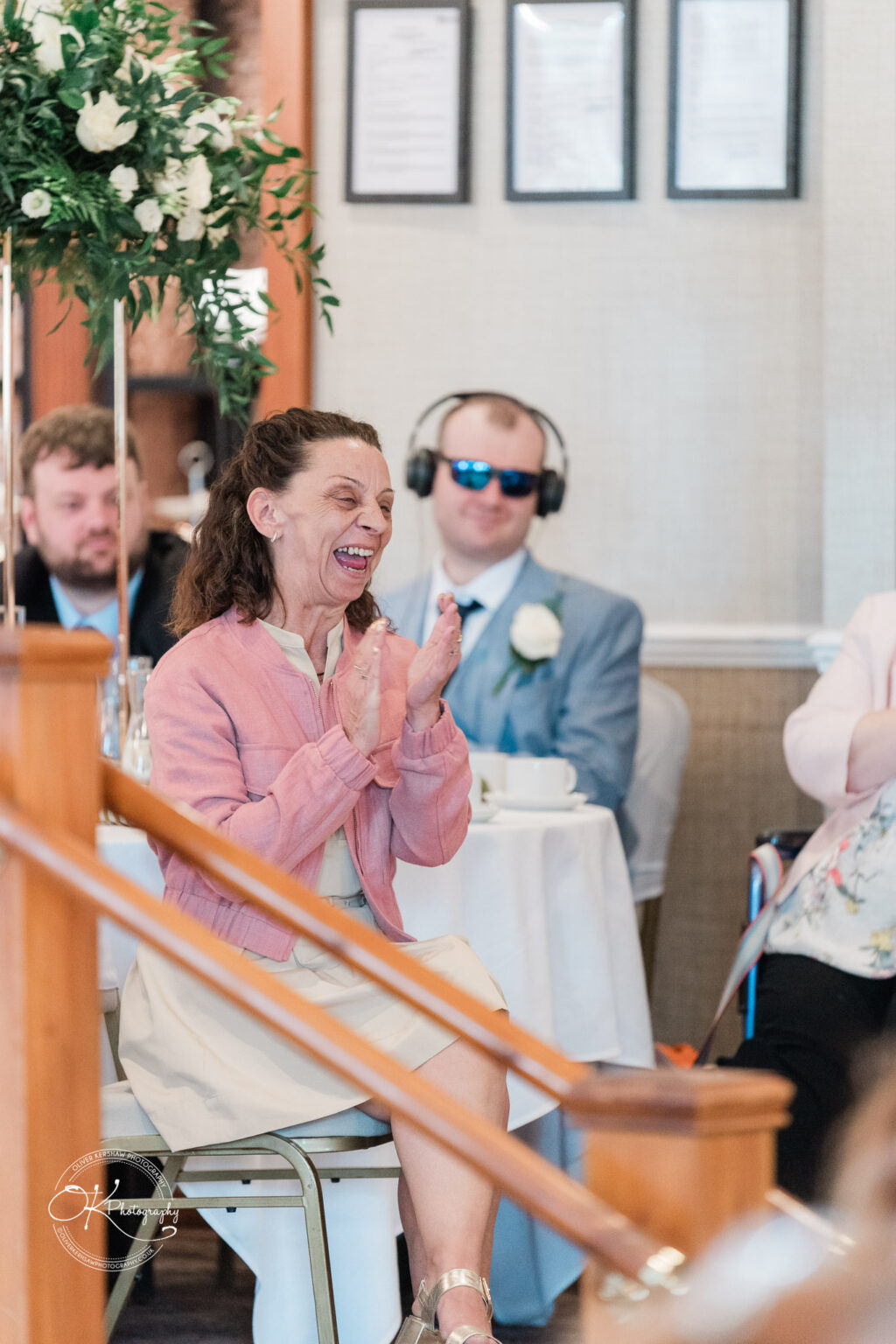 A woman in a pink jacket is sitting and laughing joyfully while clapping her hands. In the background, a man in sunglasses and headphones is seated, along with other attendees at a reception. There are decorative plants and tables set up in the room.