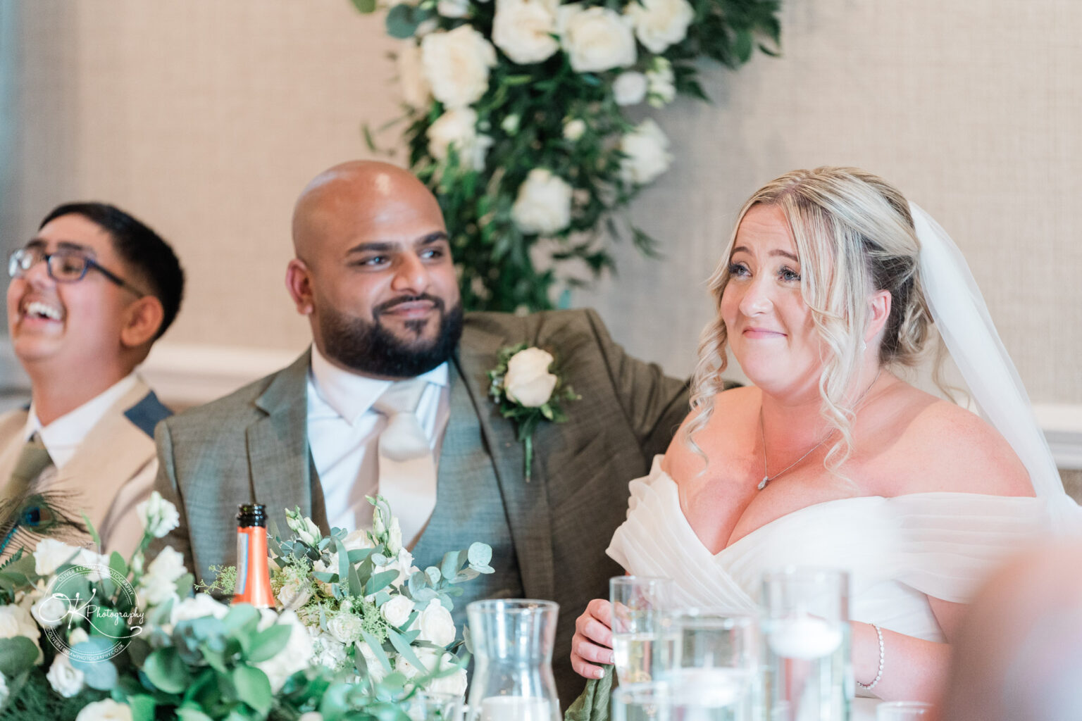 A happy bride with curly hair and a white dress sits at a table alongside a man in a suit, both smiling, while a young boy in a beige suit laughs in the background. The table is decorated with flowers and drinks.