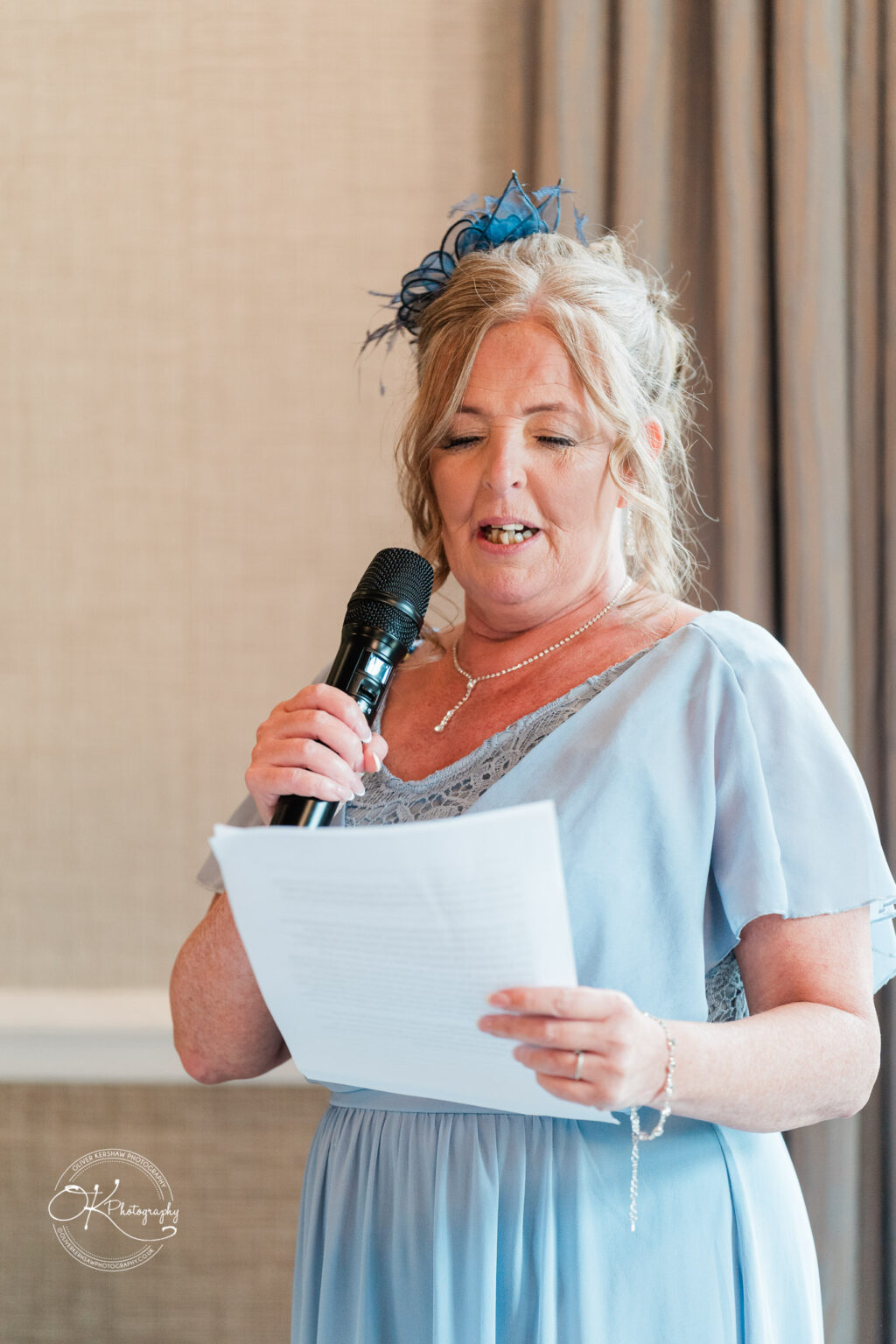 A woman in a light blue dress and matching accessories is speaking into a microphone while holding a sheet of paper, with a soft background featuring curtains.
