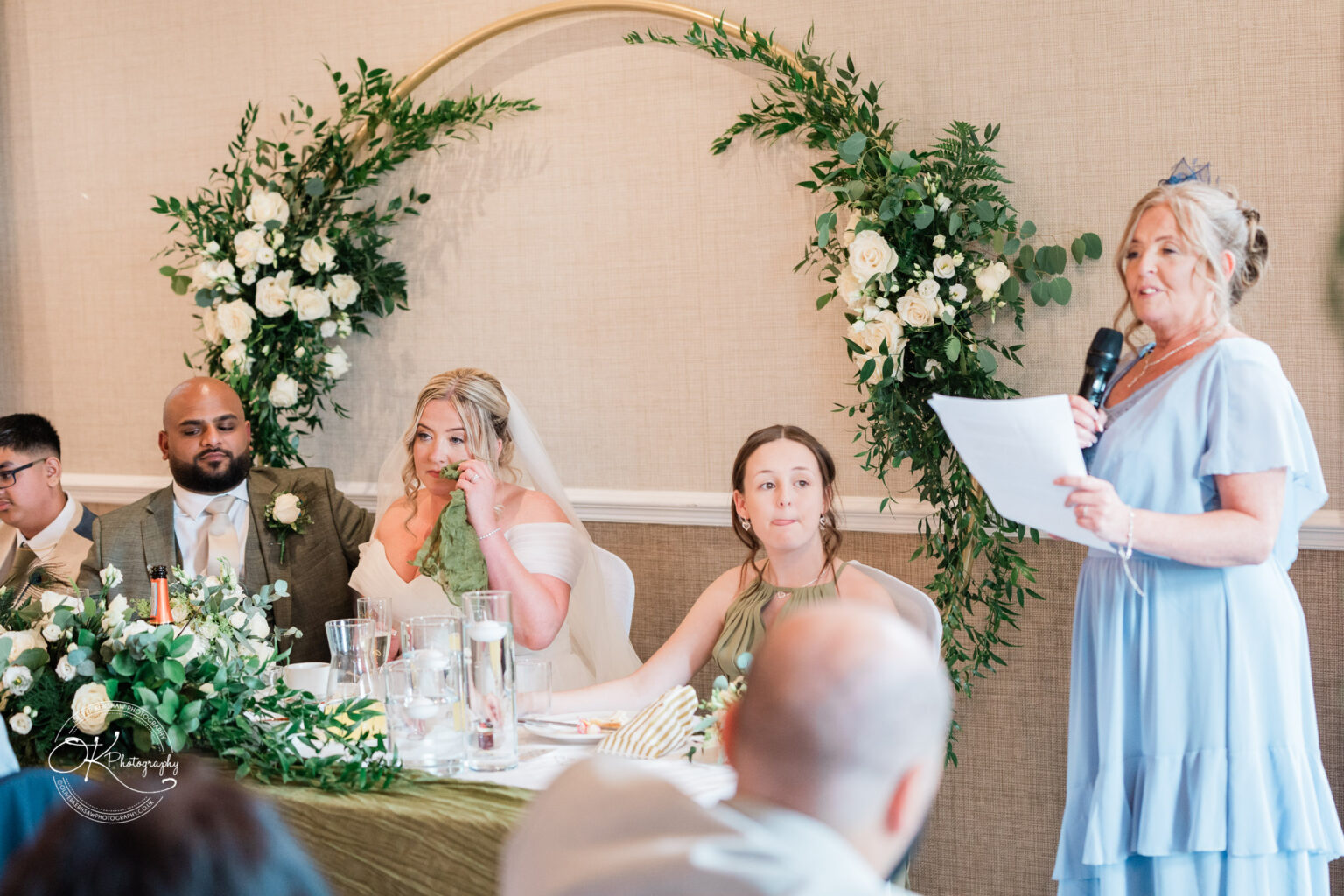 A bride seated at a table, wearing a white wedding dress and veil, is eating while a woman in a light blue dress speaks into a microphone. Another woman in a green dress listens, and a man with a beard sits beside the bride. The table is adorned with floral arrangements and glassware. In the background, a floral arch is visible.