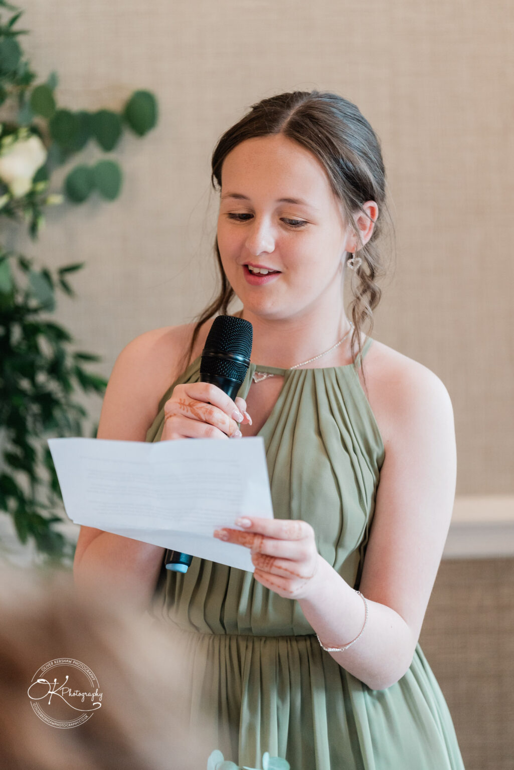 A young woman in a green dress is standing and speaking into a microphone while reading from a paper, with a friendly smile. She has long, wavy hair and is wearing simple jewelry. Blurred greenery is visible in the background.