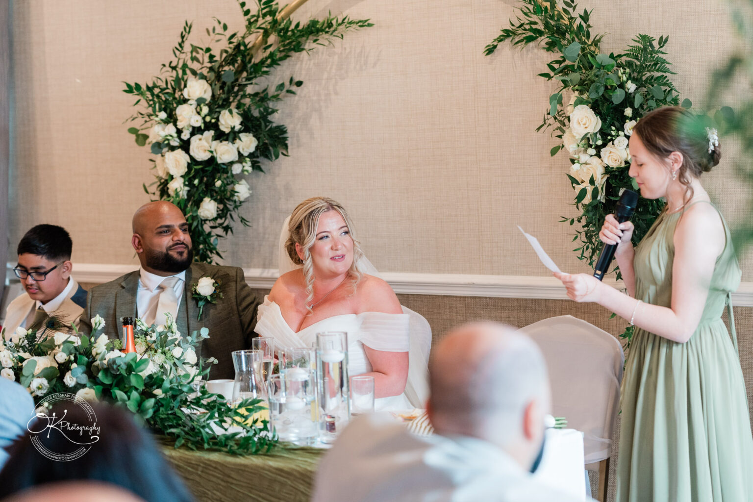 A wedding reception scene featuring a bride in a white dress and a groom in a suit seated at a decorated table, while a young woman in a green dress delivers a speech. A boy in glasses sits beside the groom, and floral arrangements are visible in the background.
